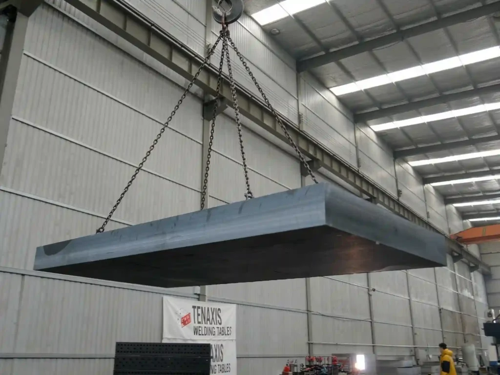 A heavy-duty steel welding table being moved by an industrial crane within the Tenaxis precision welding table manufacturing facility.