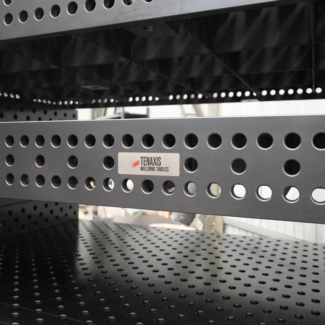 Stacked Tenaxis modular welding tables showcasing the official brand logo and precision-drilled side plates in a warehouse.