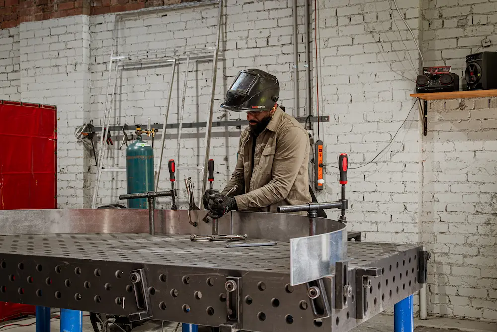 Professional welder using specialized workholding clamps and locating squares to secure a curved metal workpiece on a heavy-duty modular 3D welding table.