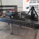 Side-angle view of a 1200x2400mm nitrided steel welding table with stainless steel adjustable legs and industrial workholding tools.
